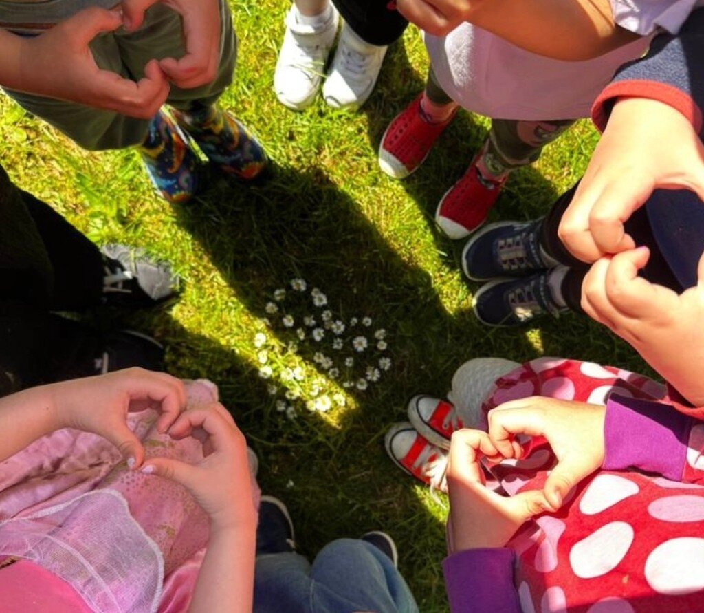 Children standing in a circle on the grass, making heart shapes with their hands around a heart of flowers